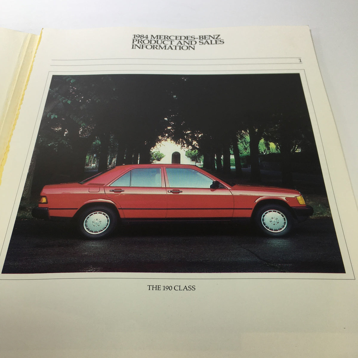 Page from the 1984 Mercedes-Benz 190-Class Product and Sales Information Catalog displaying a side view of a red Mercedes vehicle in a scenic setting.
