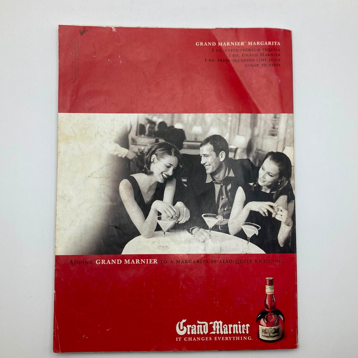 Cover of a vintage Grand Marnier advertisement featuring three people enjoying cocktails, highlighted by the tagline 'Adding Grand Marnier to a Margarita is also quite enticing.'