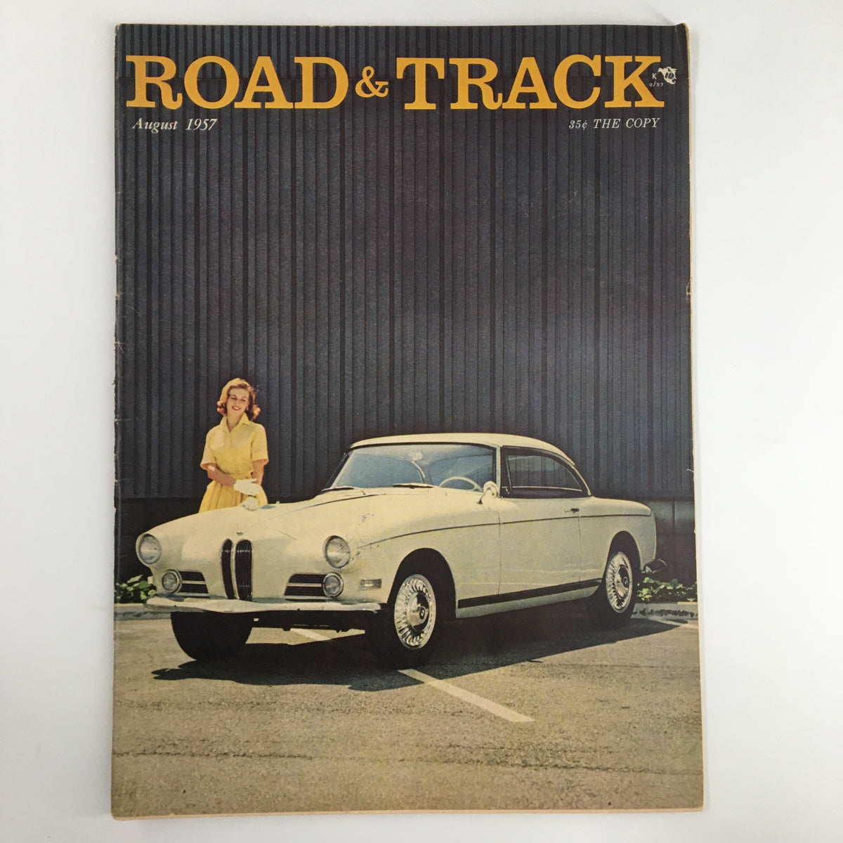 Cover of VTG Road & Track Magazine from August 1957 featuring a stylish white car and a smiling woman in a yellow dress, highlighting Jaguar XK-SSS road test.