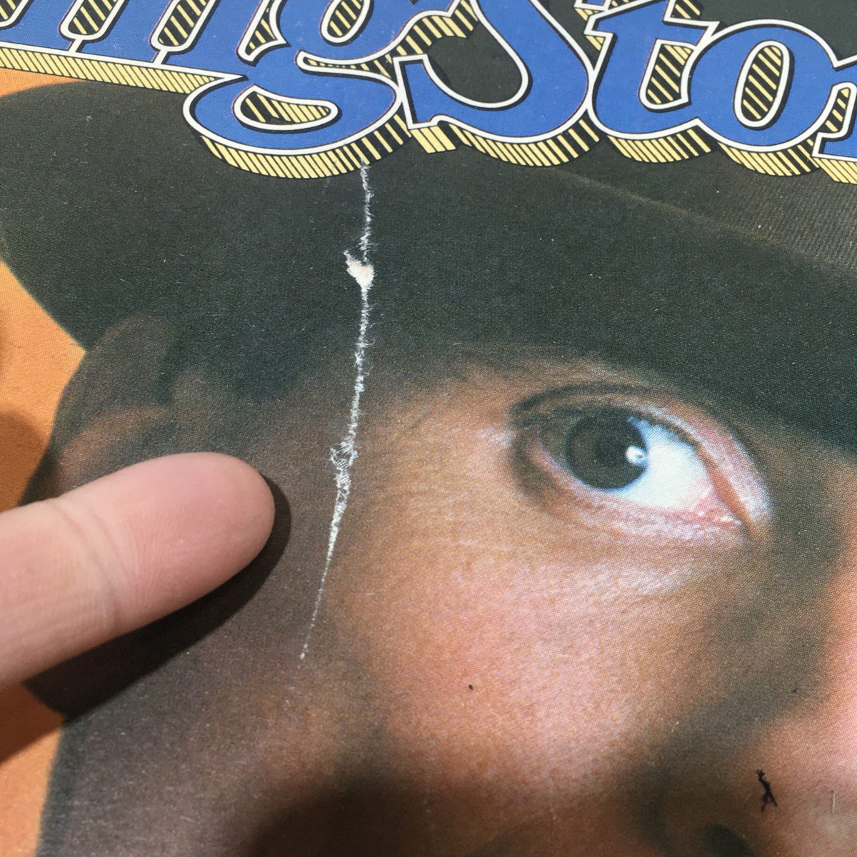 Detailed view of the front cover of a Vintage Rolling Stone magazine featuring Men At Work, highlighting a significant crack and finger pointing at the damaged area.