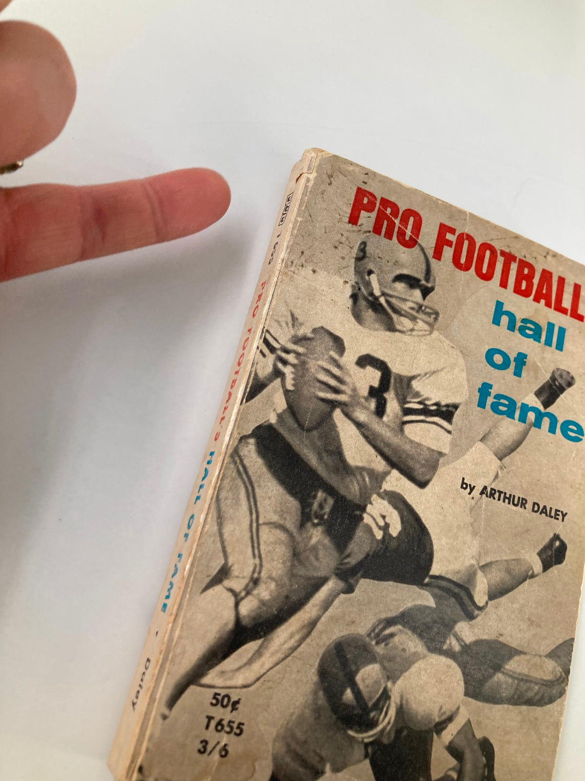 Close-up view of the Vintage 1963 Pro Football's Hall of Fame book showing the cover and the author, Arthur Daley, alongside a hand gesturing towards it.