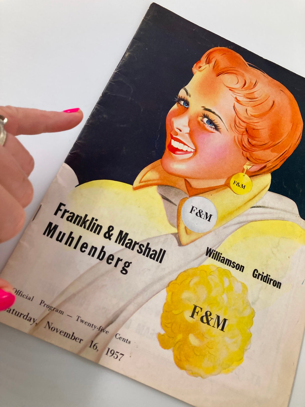 Close-up of the November 16 1957 Football Franklin & Marshal Muhlenberg Official Program cover, highlighting a smiling woman with orange hair and an F&M earring, pointing finger in view.