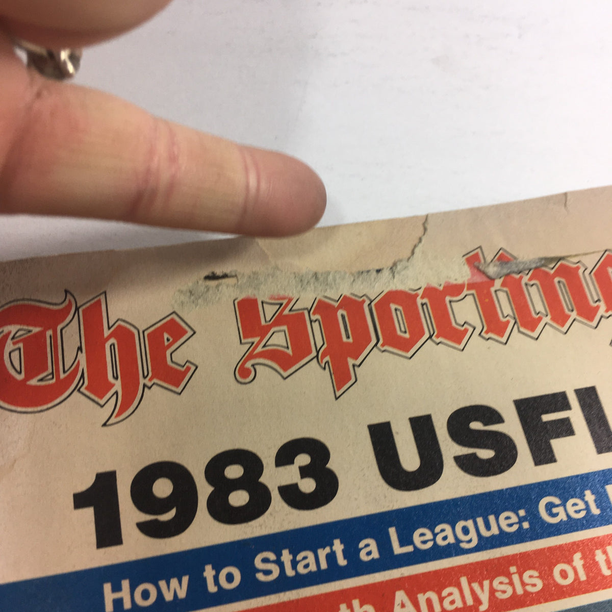 Close-up of the top edge of the VTG USFL Kickoff Official Magazine 1983, published by The Sporting News, showcasing wear and tear, including a torn area and marked text.