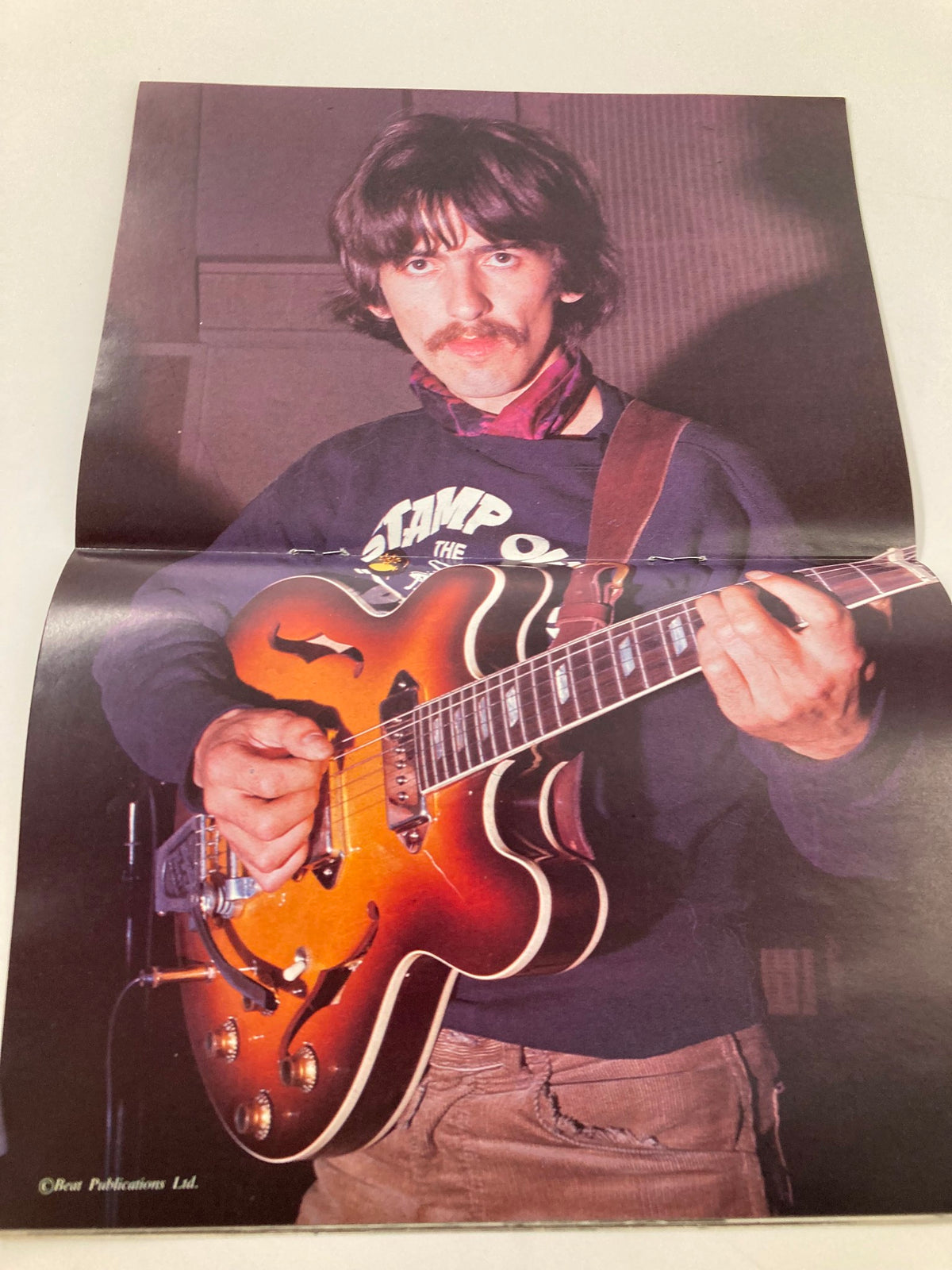 Photograph of George Harrison from The Beatles, holding an electric guitar in a casual setting. This image is part of The Beatles Book Monthly Magazine, April 1984, Issue #96, and highlights his iconic musical style.