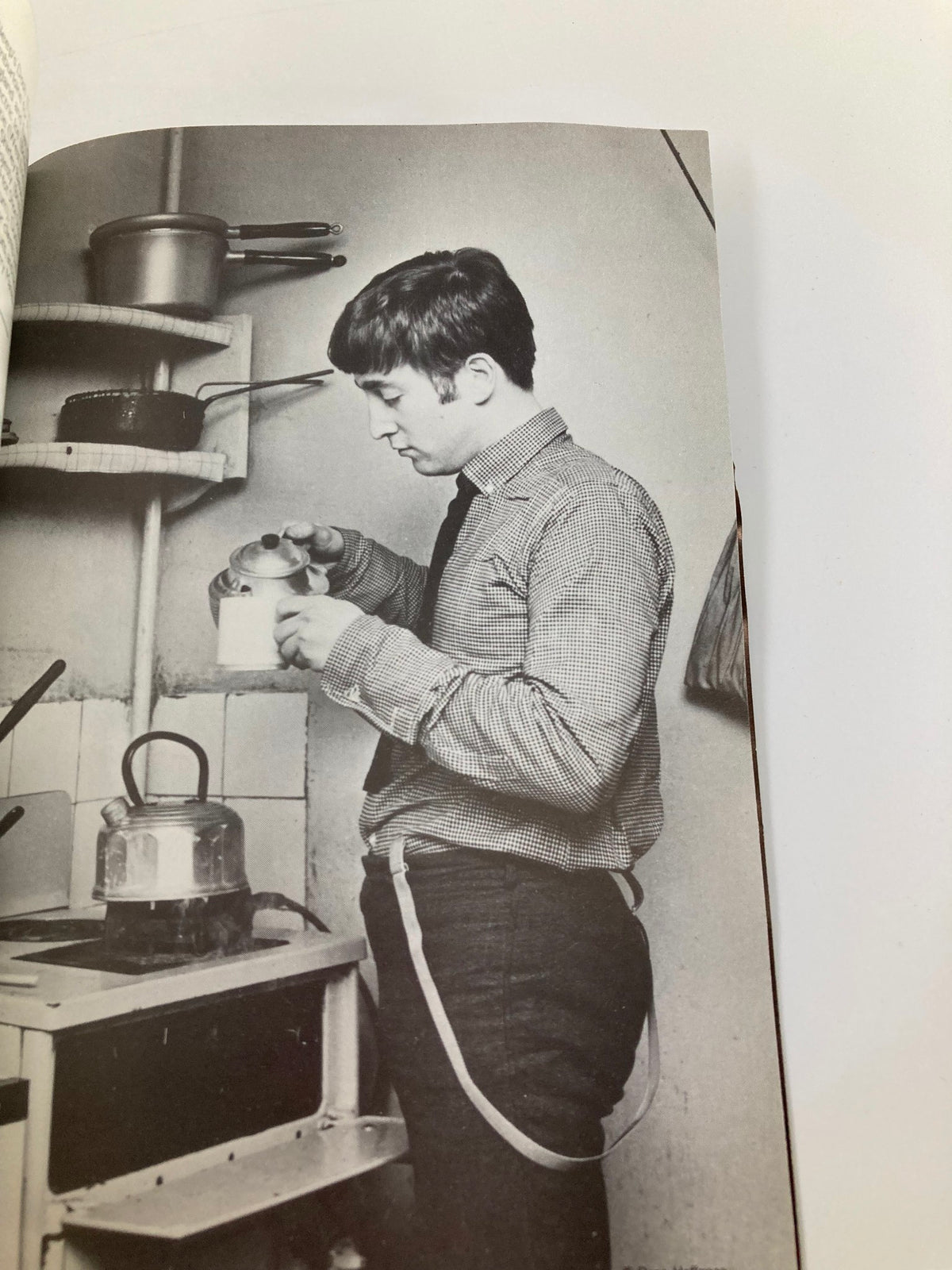 Black and white photograph of John Lennon preparing tea in a kitchen, from the 1985 John Lennon at the Astoria Theatre by Bob Eaton.