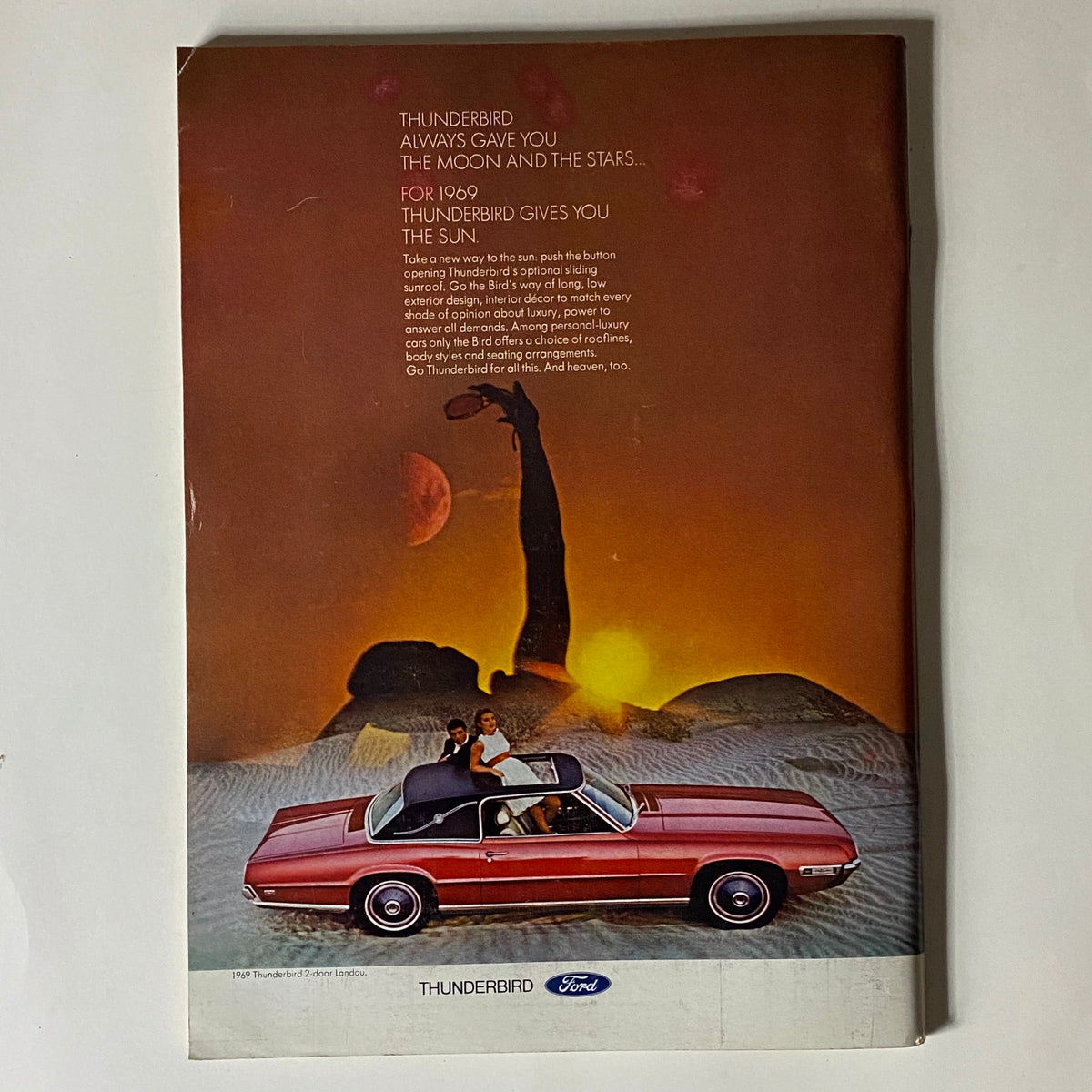 Vintage advertisement featuring a 1969 Ford Thunderbird car with a couple enjoying a scenic sunset. The ad reads "Thunderbird always gave you the moon and the stars... for 1969 Thunderbird gives you the sun." This ad highlights the luxurious features of the Thunderbird, making it a classic collectible item for enthusiasts.