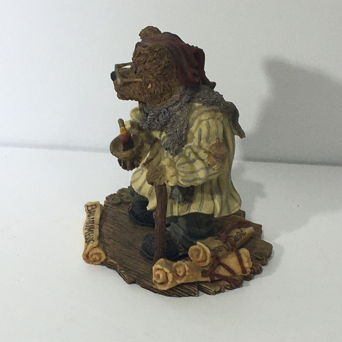 Side view of The Bearstone Collection: Style #228333 PAW Scrooge McBear, a bear figurine in a striped nightgown, holding a candle and walking stick, with details of rolled parchment and 'Bah Humbug!' sign.