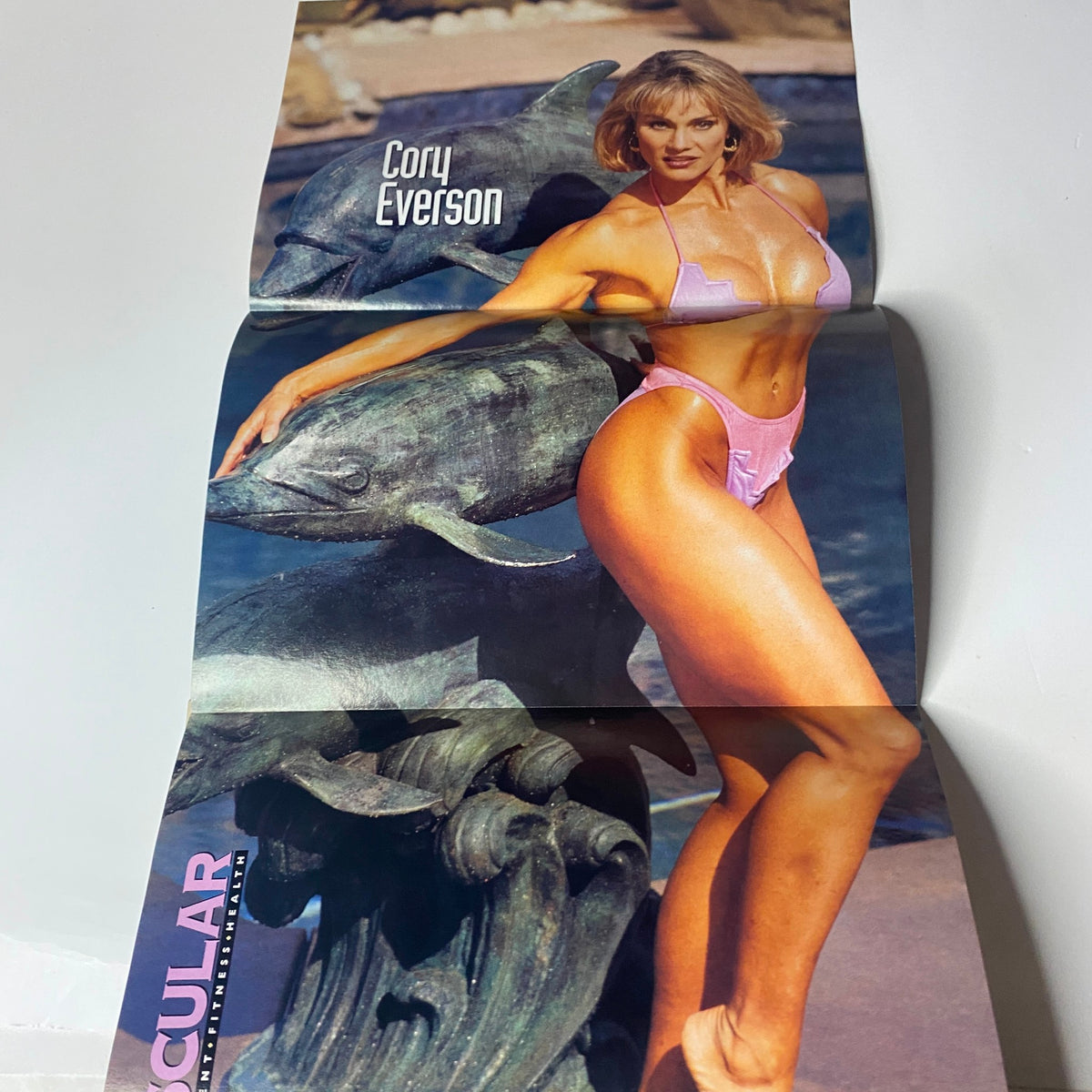 Cory Everson featured in Muscular Development Magazine April 1995, posing in a pink bikini next to a decorative statue, striking a fitness pose highlight her sculpted physique.