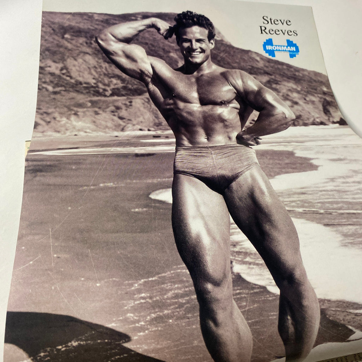 Image of bodybuilding legend Steve Reeves showcasing his physique on the beach, featured in IronMan Magazine May 1995.