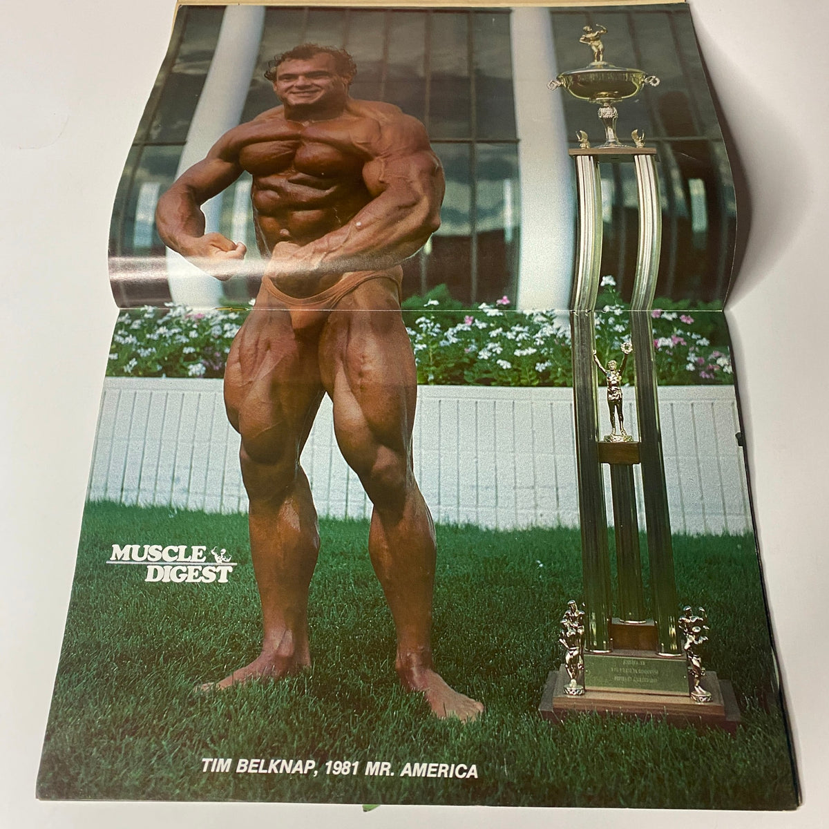 Centerfold of Muscle Digest Magazine December 1981 showcasing Tim Belknap, the 1981 Mr. America, posing next to his trophy in an outdoor setting.