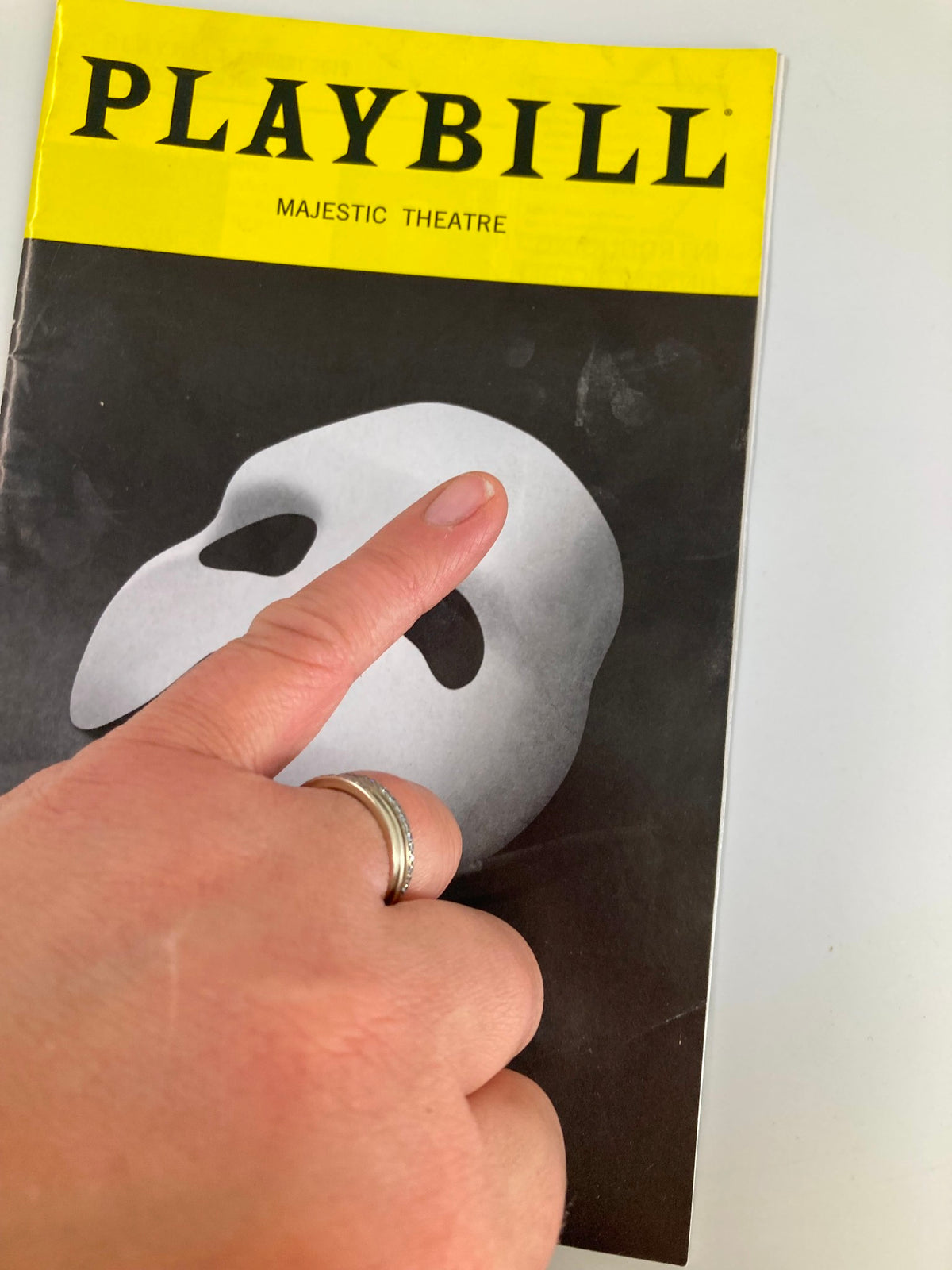 Close-up of the 2019 Playbill for The Phantom of the Opera at Majestic Theatre, with a hand pointing at the Phantom mask design.