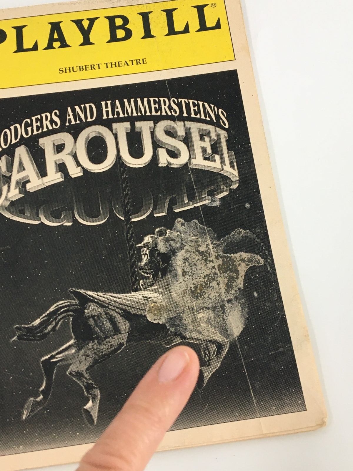 Close-up view of the front cover of the 1996 Playbill for Rodgers and Hammerstein's Carousel, highlighting the horse and carousel graphic along with a finger pointing to a specific area.