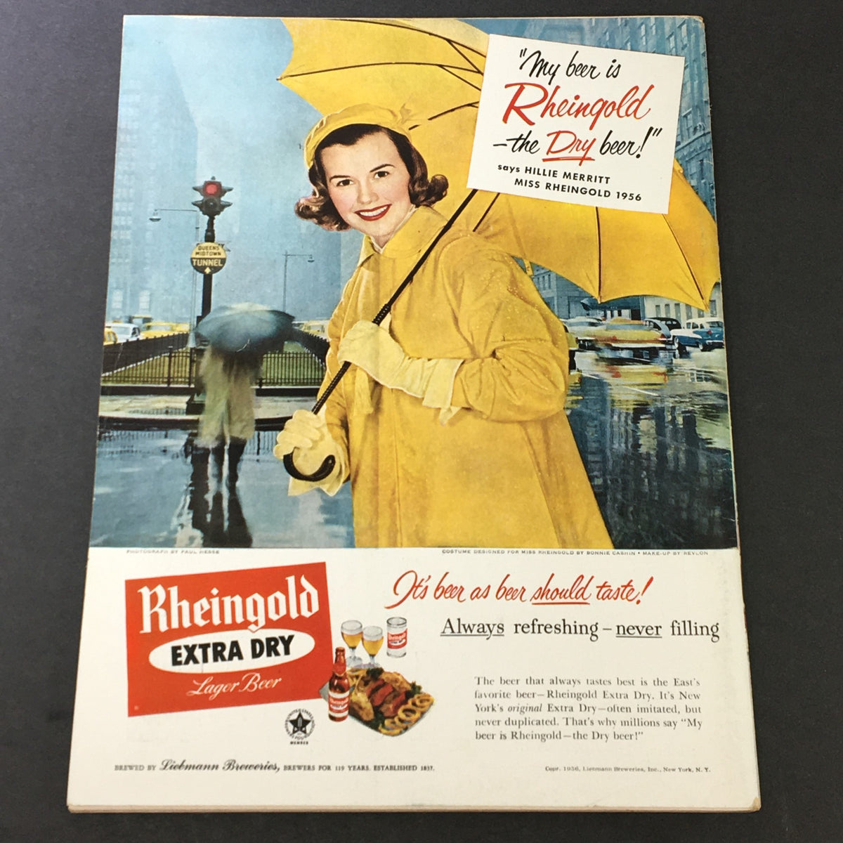 Advertisement page from VTG Gourmet The Magazine of Good Living March 1956 showing a woman in a yellow raincoat under an umbrella promoting Rheingold Beer, emphasizing mid-century design and lifestyle.