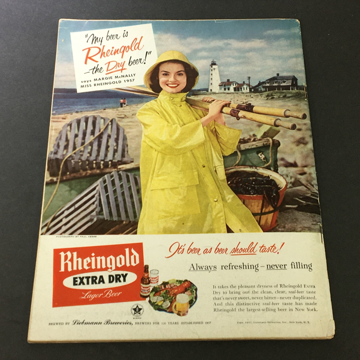 Vintage advertisement for Rheingold Extra Dry Lager Beer featuring a woman dressed in yellow rain gear, promoting the beer with a coastal background, from Miss Rheingold 1957.