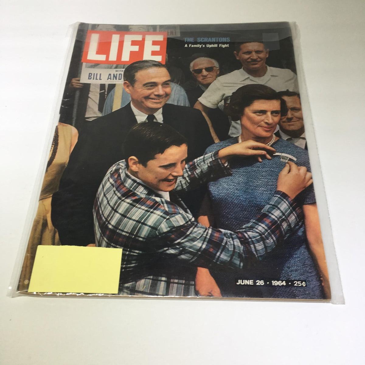 Cover of Life Magazine from June 26, 1964, featuring 'The Scrantons: A Family's Uphill Fight.' The image shows a joyful young boy in a plaid shirt assisting a woman with a name tag, while others look on in the background, capturing a moment from American history.