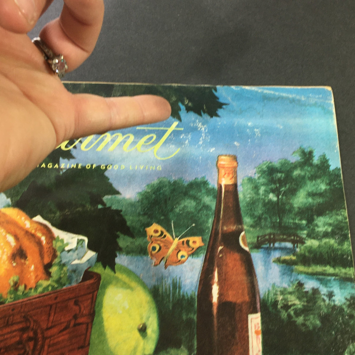 Close-up view of the cover of VTG Gourmet: The Magazine of Good Living from July 1959, highlighting details like a butterfly and a bottle near a picturesque landscape.