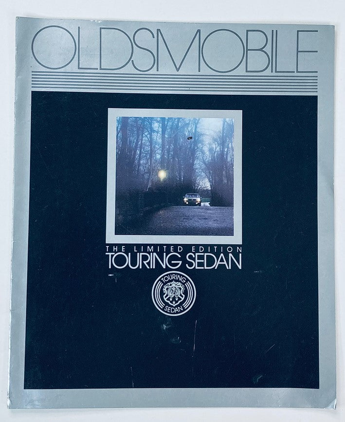 Cover of the 1987 Oldsmobile Touring Sedan Dealer Showroom Sales Brochure Guide Catalog featuring a vintage car and a misty forest background.