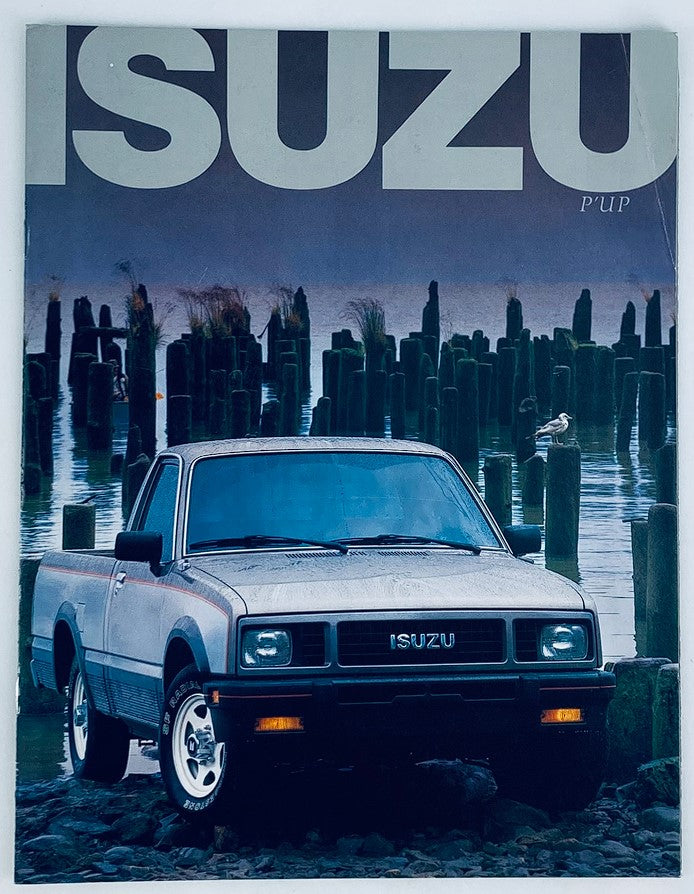 1987 Isuzu Pickup Lineup Dealer Showroom Sales Brochure featuring a front view of a silver Isuzu truck parked near water with wooden posts in the background, highlighting the vehicle's design and outdoor setting.