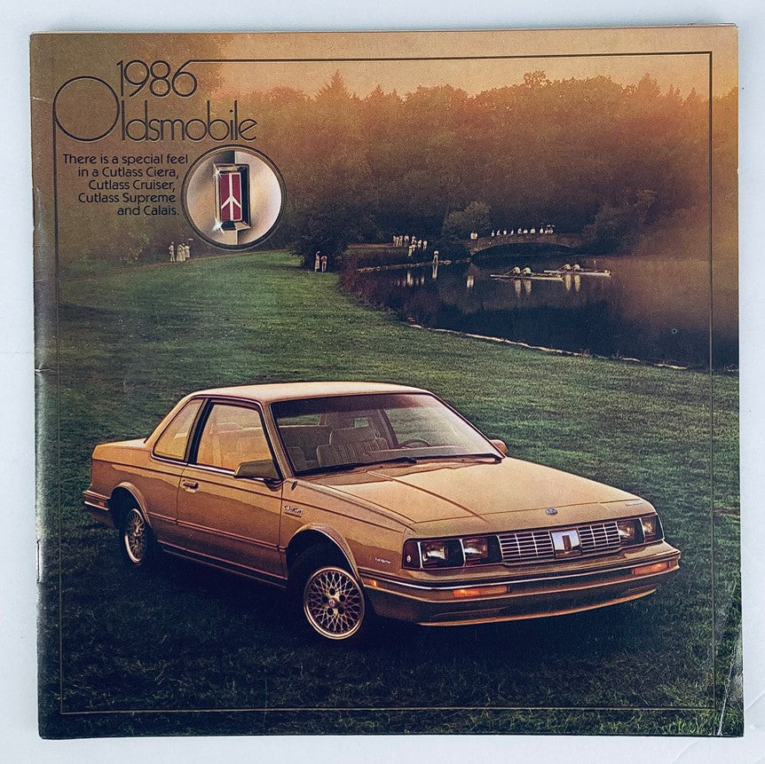 Cover of the 1986 Oldsmobile Cutlass Ciera Dealer Showroom Sales Brochure Guide Catalog featuring a gold Oldsmobile Cutlass parked on a grassy area near a serene lake at sunset.