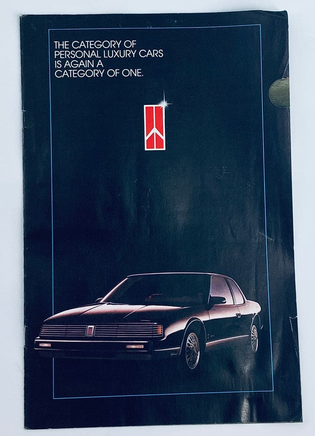 Cover of the 1986 Oldsmobile Lineup Dealer Showroom Sales Brochure showcasing a black luxury car with a distinctive logo, emphasizing the category of personal luxury cars.