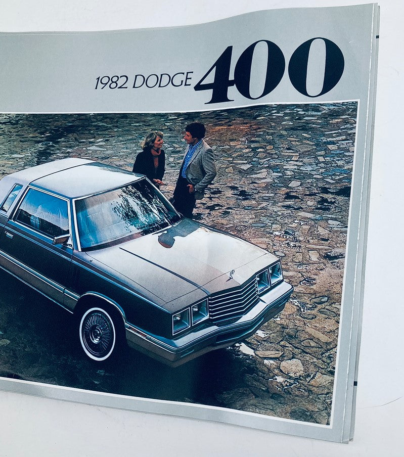 Close-up of the 1982 Dodge 400 Dealer Showroom Sales Brochure Guide Catalog showcasing a gray Dodge 400 parked on cobblestones, with two figures engaged in conversation.